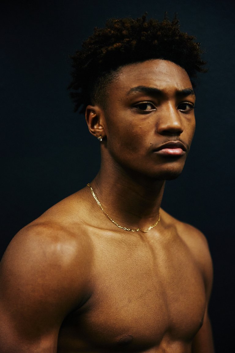 Portrait of an African American man with short dark hair and a gold chain against a dark background. Focused on upper body and face.