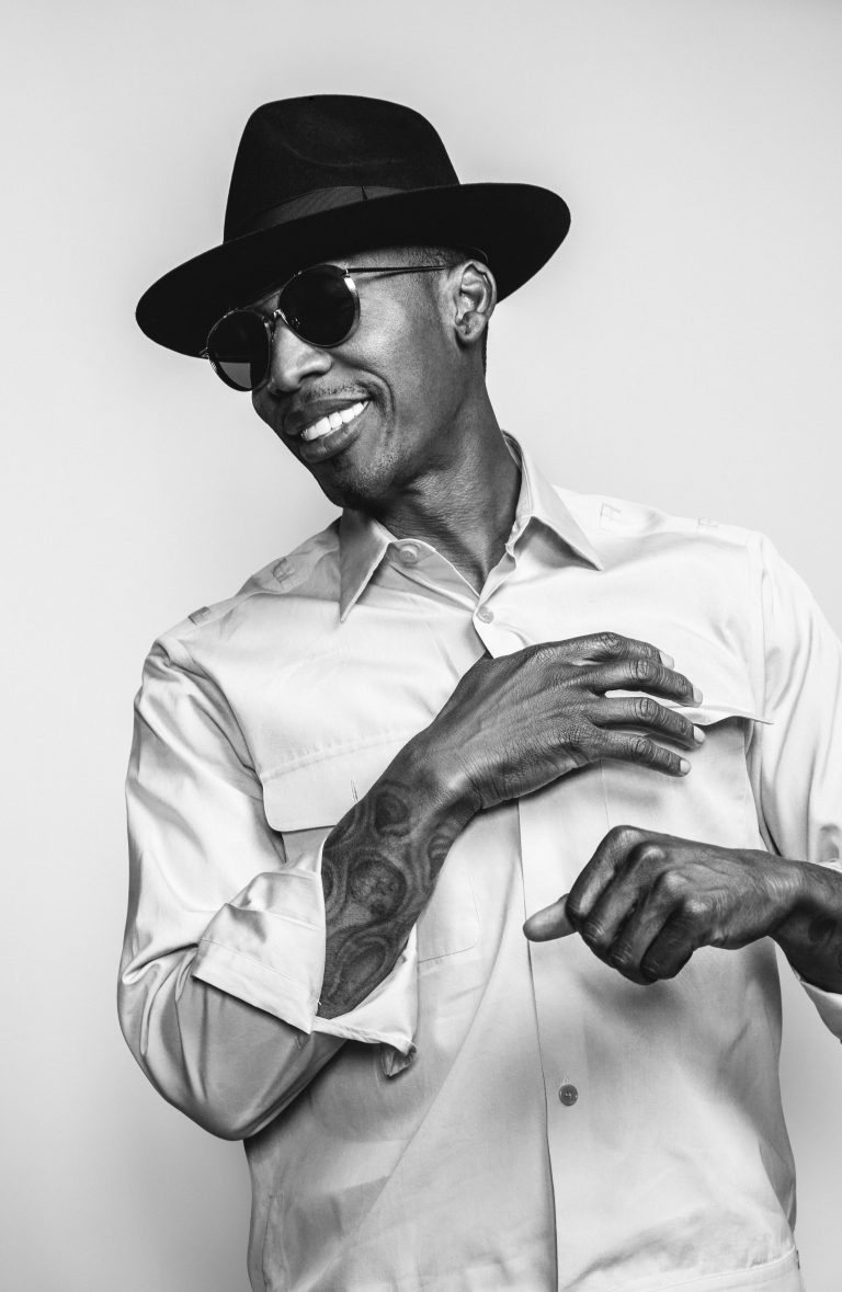 Black and white portrait of a smiling man in a fedora and sunglasses wearing a button-down shirt, photographed by G L Askew Ii.