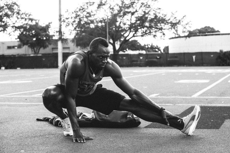 An athletic adult male in sportswear stretching his leg on a black sports track outdoors on a sunny day. Trees and a building in background.