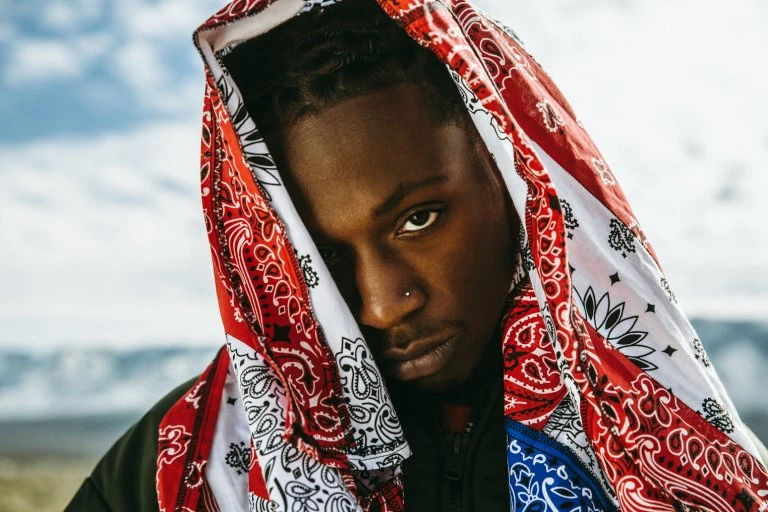 Close-up portrait of a young Black man wearing a patterned bandana as a head covering, set against a background of a bright sky.