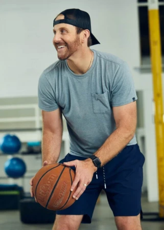 Man in sports clothes smiles while holding a basketball. He is indoors in a gymnasium setting. The image has eye-level framing.