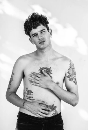 Portrait of a shirtless tattooed man with dark curly hair posing against a light background by G L Askew Ii
