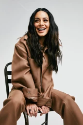 Vanessa Hudgens sitting on a chair wearing a brown jacket and trousers against a white background