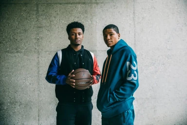 Two black men standing against a textured concrete wall, holding a basketball and wearing sportswear in Los Angeles.