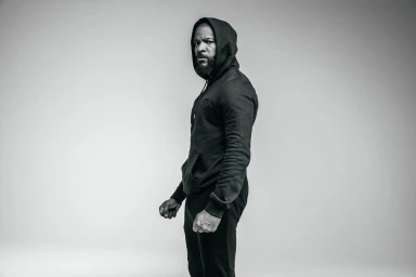 Black and white portrait of a black man wearing a dark hooded sweatshirt against a seamless studio background