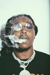 Portrait of Takeoff wearing round gold sunglasses and layered diamond necklaces while exhaling white smoke against a white backdrop.