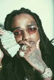 Portrait of rapper Quavo holding a thick stack of cash to his face with tattooed hands wearing diamond rings and a watch