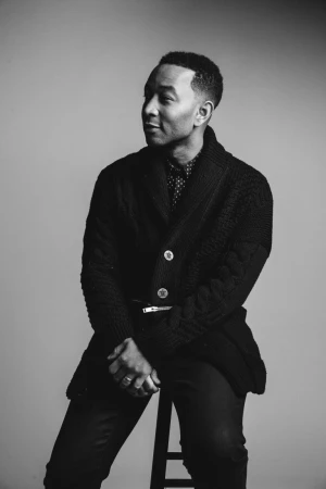 Black and white portrait of John Legend seated on a stool wearing a knitted cardigan and patterned shirt against a grey backdrop.