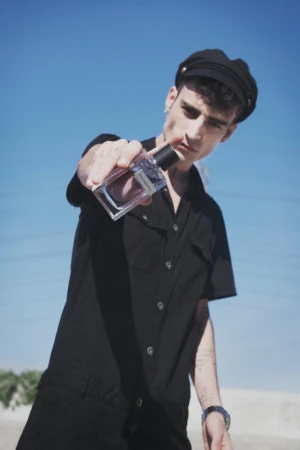 Young man wearing black cap and black shirt holding up a perfume bottle outdoors under a clear blue sky