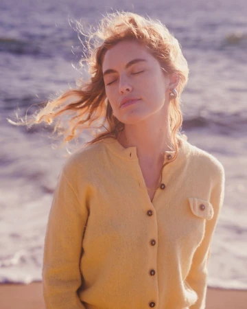 Woman in a yellow cardigan standing by the ocean with her eyes closed as wind blows her hair, photographed by Nastasia Dusapin