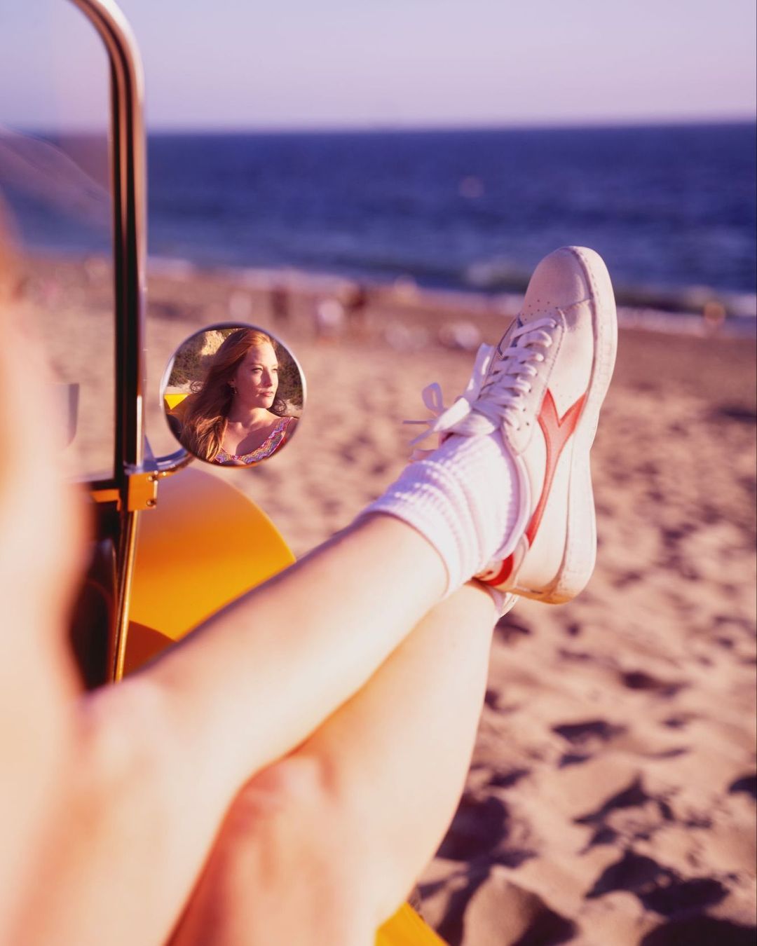 Legs up in a car on a sandy beach with a side mirror reflecting a woman and the ocean in the background on a sunny day.