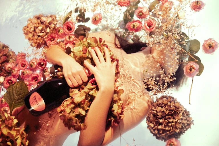 Woman lies in water covered in pink and white flowers while holding a bottle of champagne, illuminated by soft, diffused light.