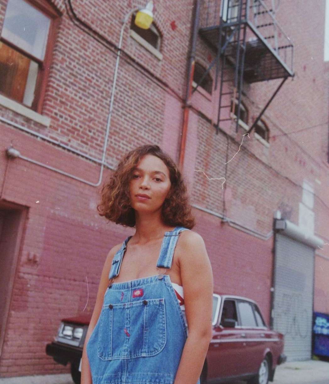 Young adult woman in denim overalls stands outside a brick building with a vintage car parked nearby in an urban setting.