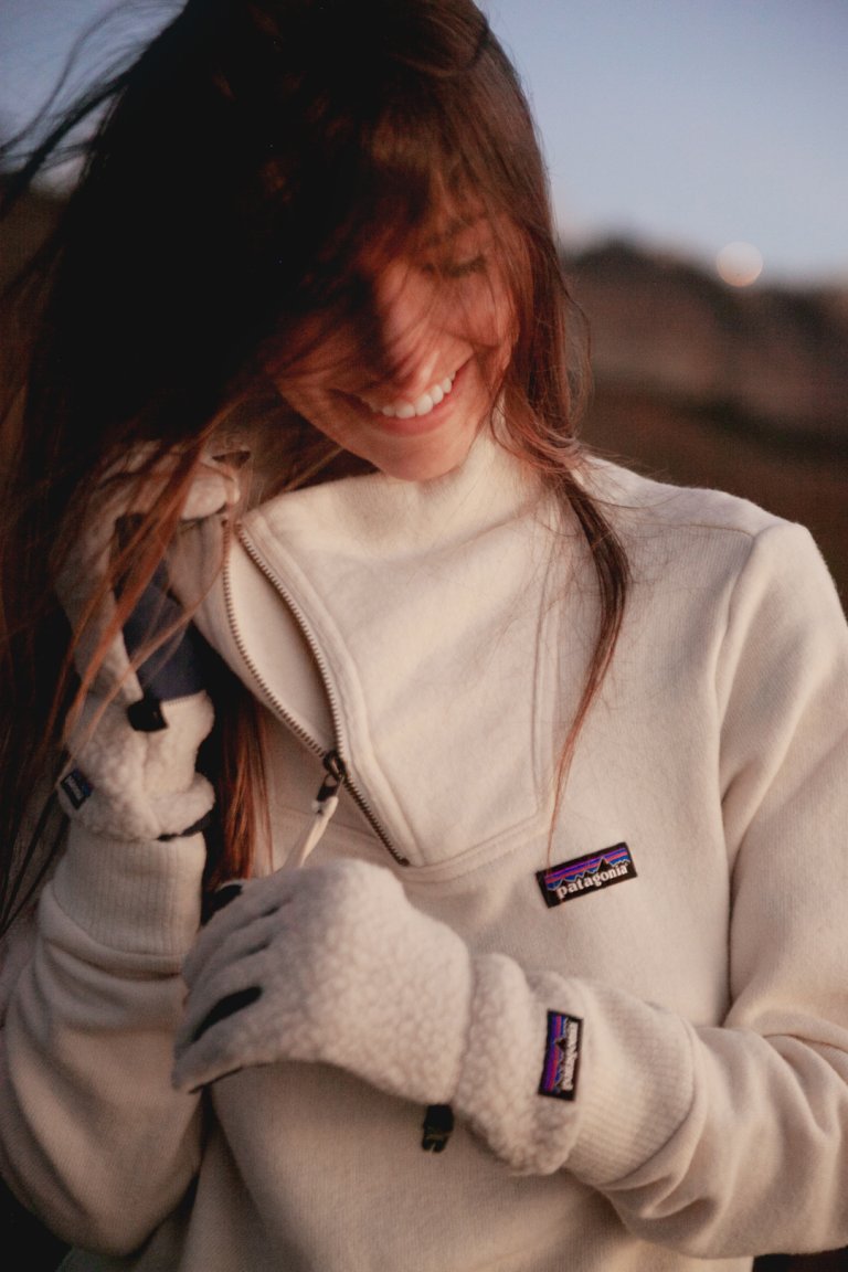 Smiling woman in outdoor setting wearing a white Patagonia fleece quarter-zip with fleece gloves and long brown hair.