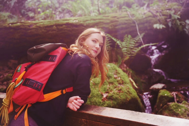 Woman with a red backpack in a forest leans on a wooden railing with a waterfall and ferns in the background, looking at the camera.