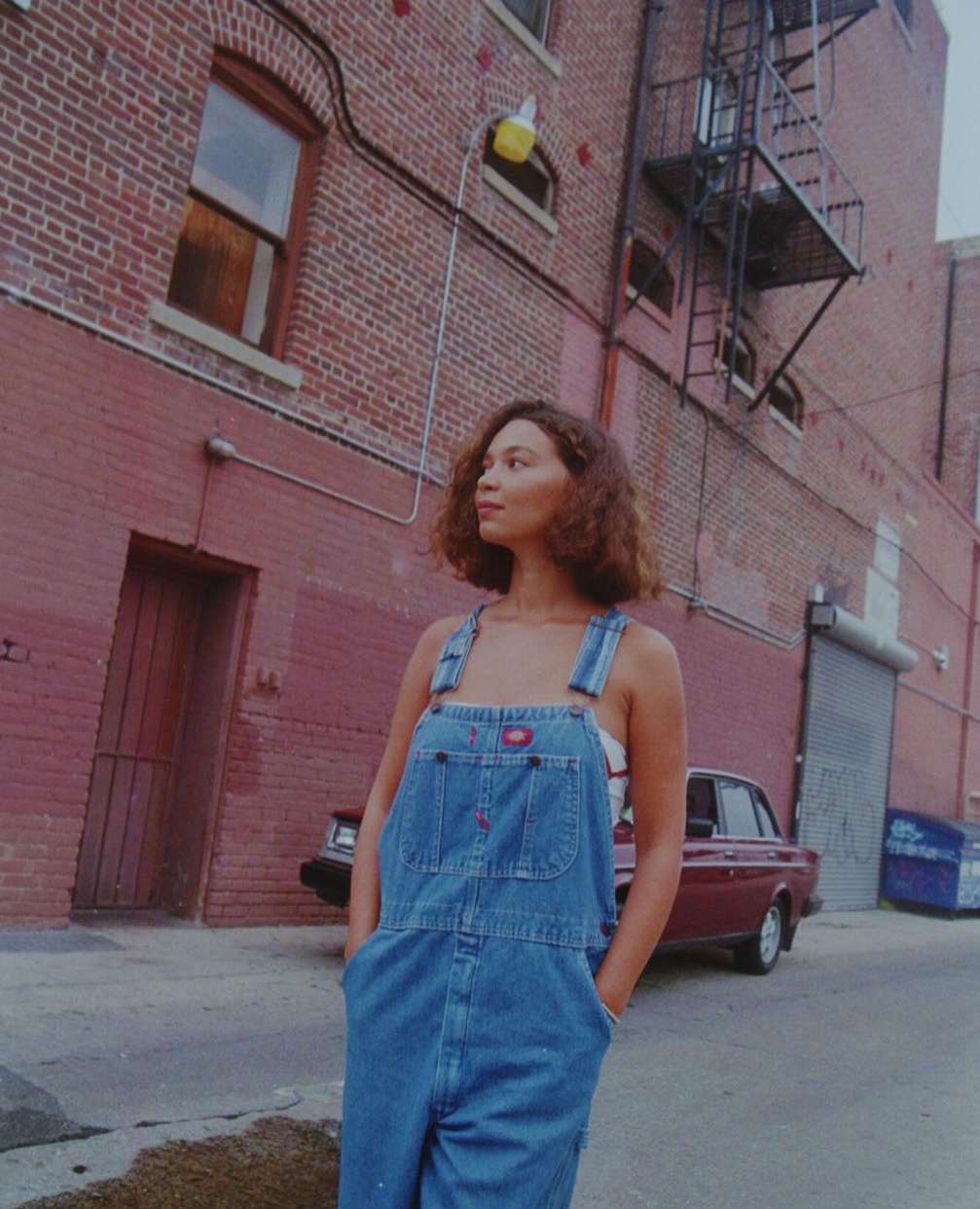 A young woman in denim overalls stands on a city street with brick buildings and a vintage car in the background on a cloudy day.