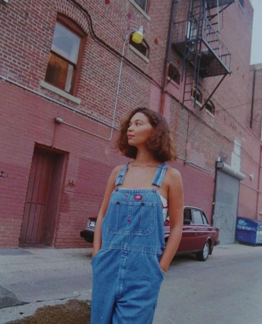 A young woman in denim overalls stands on a city street with brick buildings and a vintage car in the background on a cloudy day.