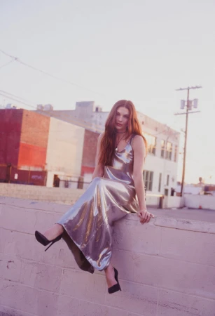 Woman in a long metallic silver slip dress seated on a concrete wall outdoors with urban buildings in the background.