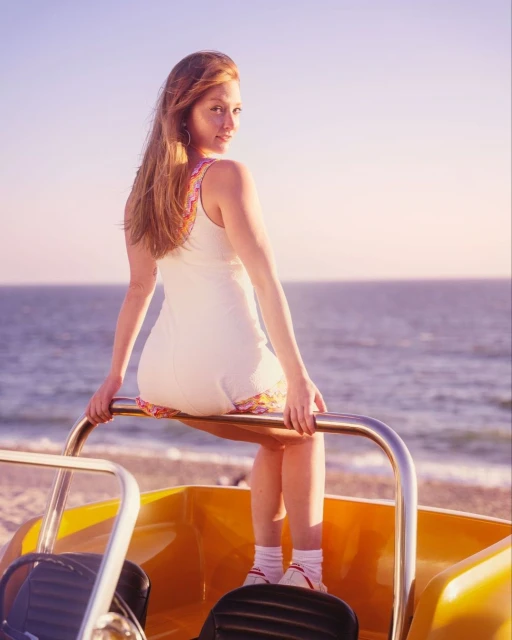 Woman in white dress sits on a yellow dune buggy on a sandy beach with ocean background, looking over her shoulder.
