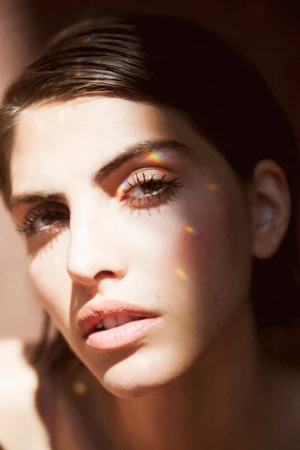 Close-up portrait of a woman with dark hair and prismatic rainbow light refractions on her face, photographed by Nastasia Dusapin.