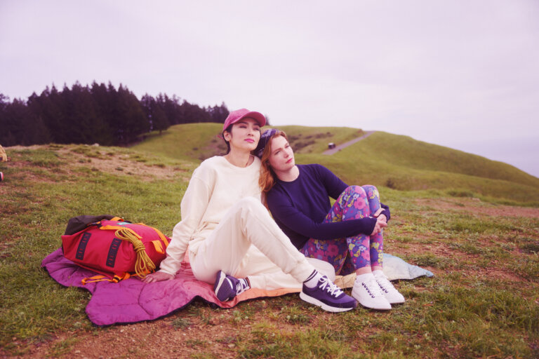 Two women sitting together on a grassy hill with a red backpack and magenta sleeping bag, trees and a path in the background.