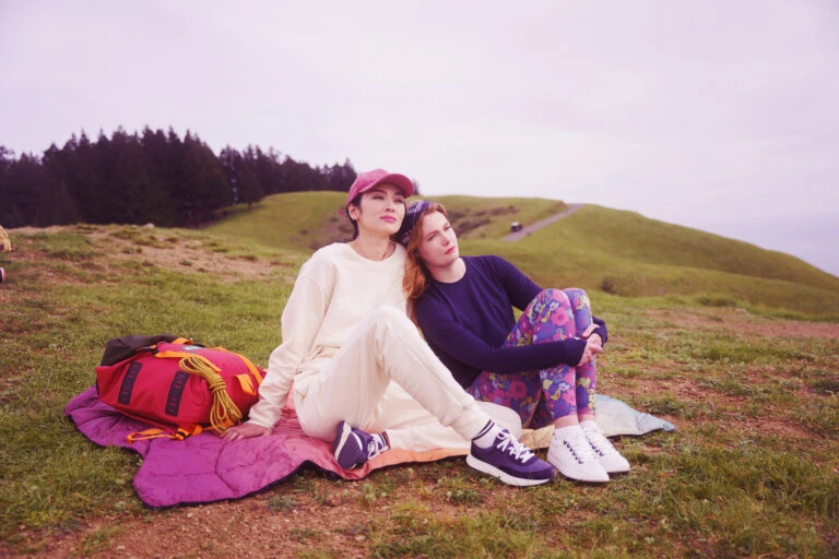 Two women sitting together on a grassy hill with a red backpack and magenta sleeping bag, trees and a path in the background.