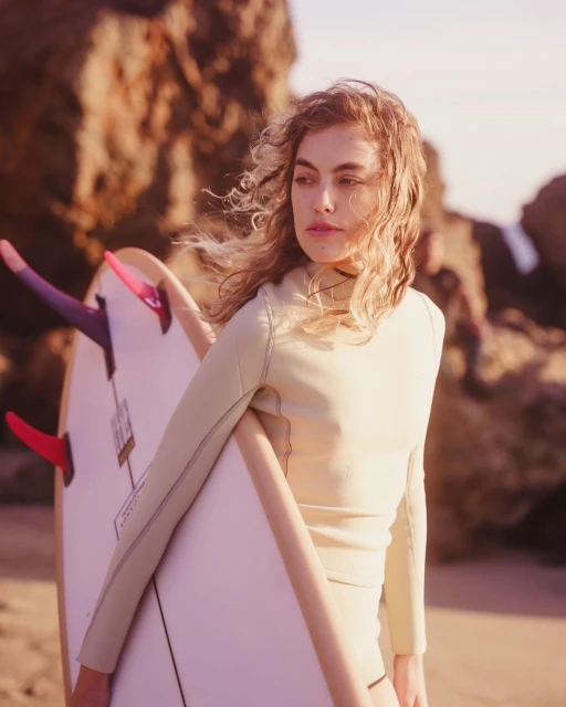 Young woman in wetsuit carrying a surfboard on a sandy beach with rocks in the background during daytime, outdoor lifestyle and sports.