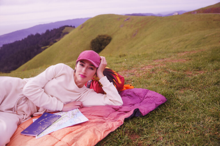 Woman in pink hat lies on orange blanket with map in grassy field with rolling hills. Portrait framing shows outdoor recreation.