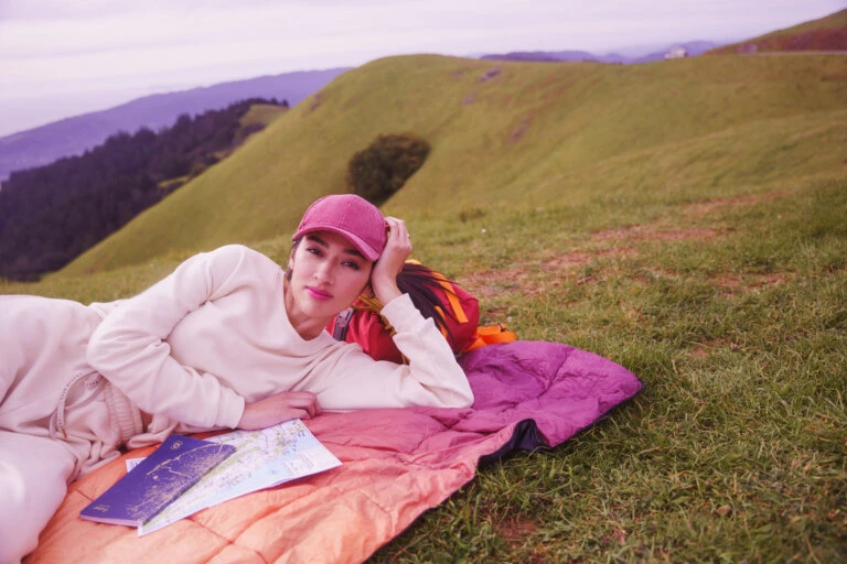 Woman in pink hat lies on orange blanket with map in grassy field with rolling hills. Portrait framing shows outdoor recreation.