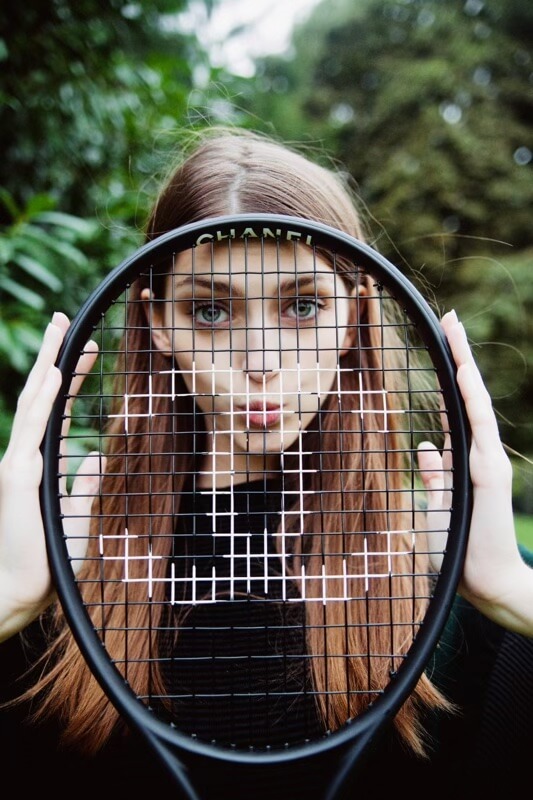A young woman with long brown hair poses outdoors holding a Chanel-branded tennis racket, with a green foliage background, portrait orientation.