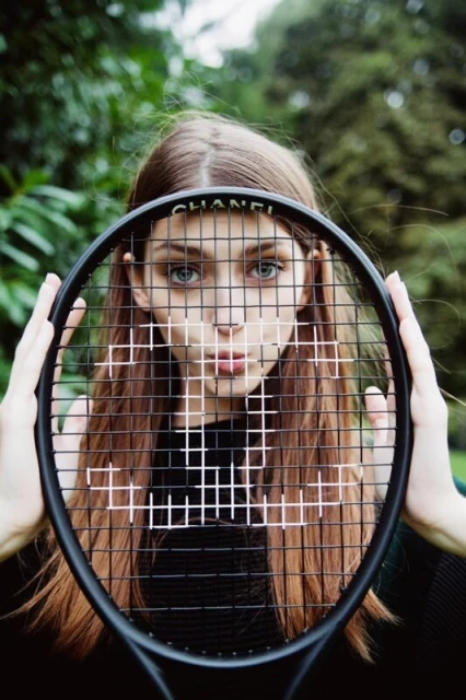 A young woman with long brown hair poses outdoors holding a Chanel-branded tennis racket, with a green foliage background, portrait orientation.