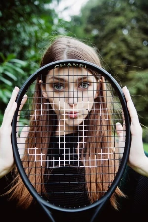 A young woman with long brown hair poses outdoors holding a Chanel-branded tennis racket, with a green foliage background, portrait orientation.