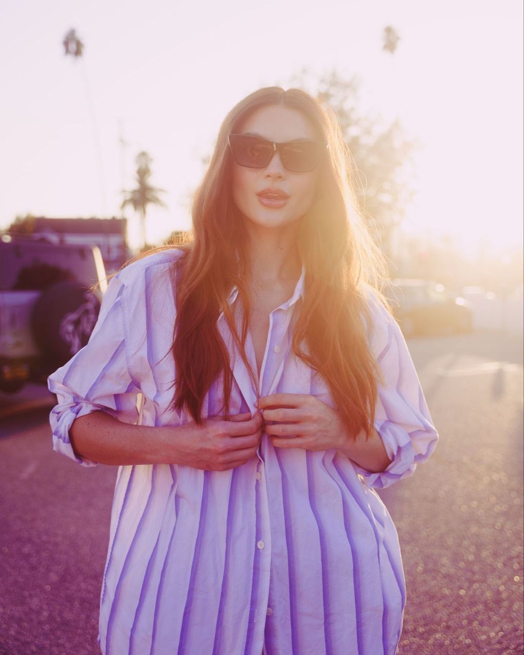 Woman in dark sunglasses wearing an oversized striped shirt outdoors against a golden sunset sky by Nastasia Dusapin