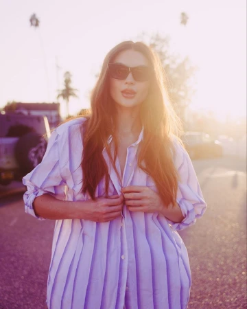 Woman in dark sunglasses wearing an oversized striped shirt outdoors against a golden sunset sky by Nastasia Dusapin