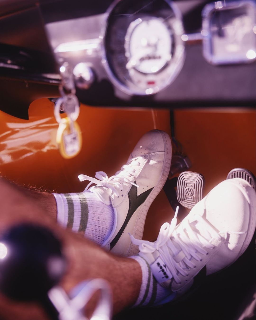 Driver's perspective of vintage car interior featuring white Diadora sneakers on pedals, green striped socks, and an orange dashboard.