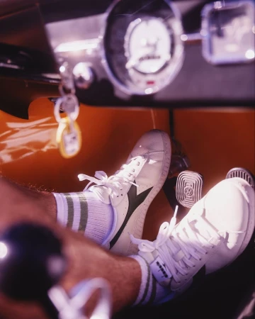 Driver's perspective of vintage car interior featuring white Diadora sneakers on pedals, green striped socks, and an orange dashboard.