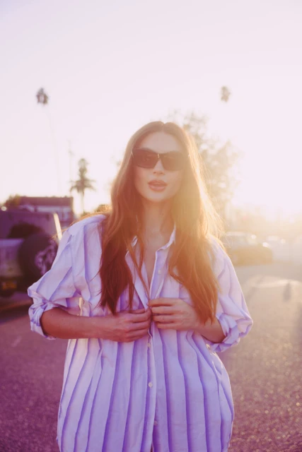 Adult woman in sunglasses and an oversized striped shirt stands on a street with cars and palm trees in the background on a sunny day.