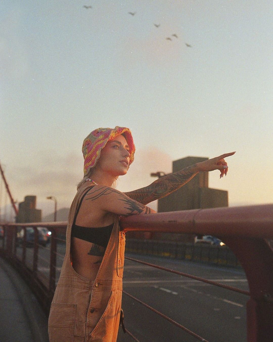 Tattooed woman in overalls and colorful hat points at birds over the Golden Gate Bridge during the daytime with neutral color grading.
