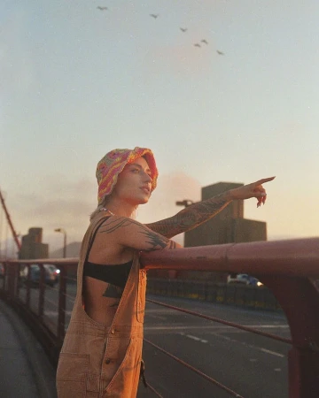 Tattooed woman in overalls and colorful hat points at birds over the Golden Gate Bridge during the daytime with neutral color grading.