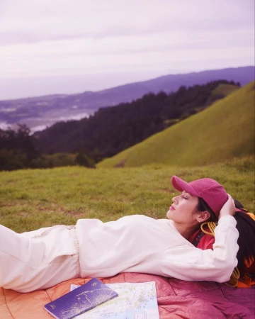 Woman wearing a pink cap and cream sweatshirt resting on a blanket in a grassy field with rolling hills and distant water.