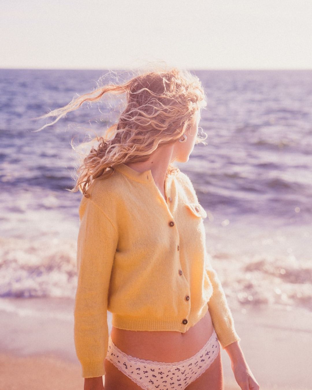 Woman with curly blonde hair wearing a yellow cardigan standing by the ocean on a sandy beach