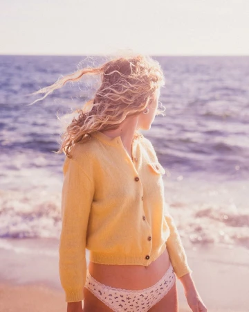 Woman with curly blonde hair wearing a yellow cardigan standing by the ocean on a sandy beach