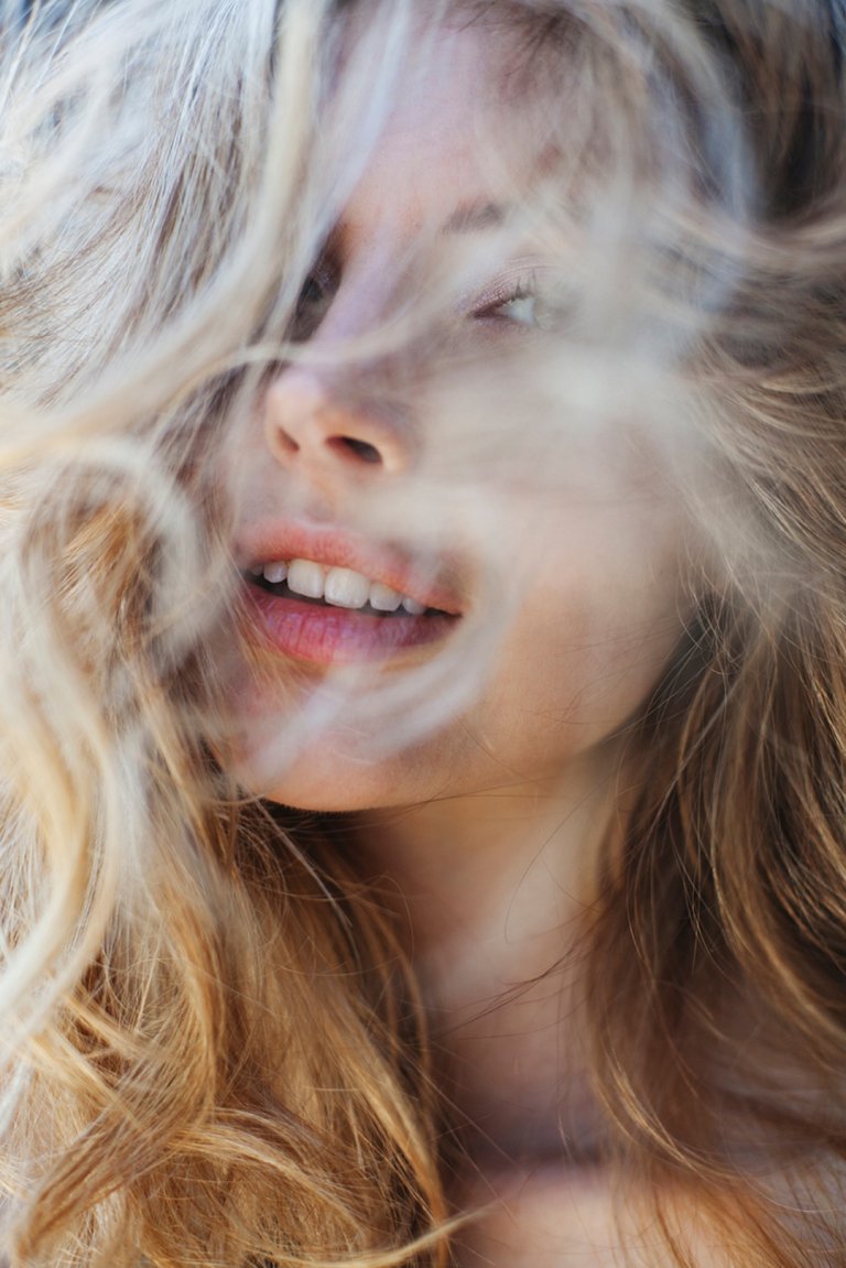 Close-up portrait of a fair-skinned adult woman with blond hair partially covering her face, and an open mouth with visible teeth.