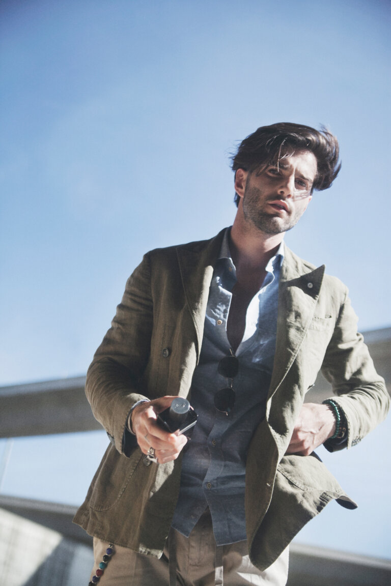 A man in an olive blazer and blue shirt holds a small black box, posing outdoors with a clear sky and urban structure backdrop.