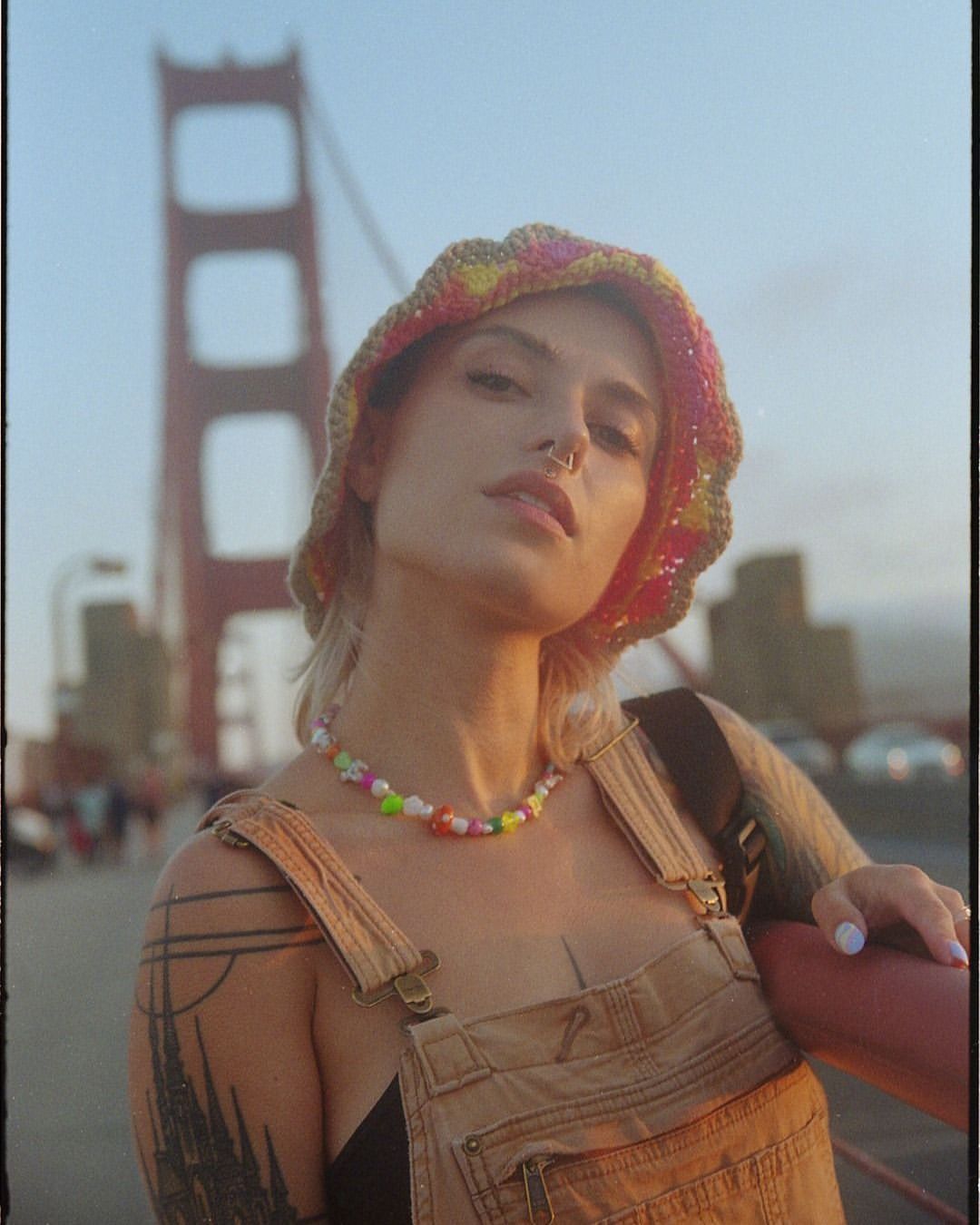 Woman poses on the Golden Gate Bridge wearing a crocheted hat and beaded necklace on a sunny day with a blurred background.