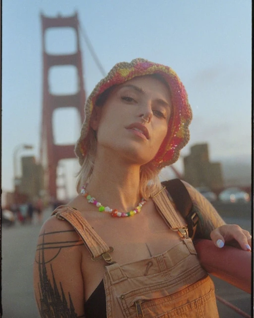 Woman poses on the Golden Gate Bridge wearing a crocheted hat and beaded necklace on a sunny day with a blurred background.