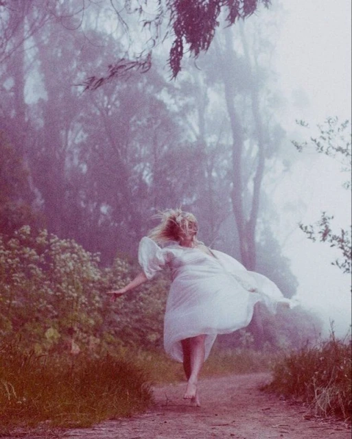 Woman in white dress runs barefoot through a misty forest, trees and greenery in background, creating an ethereal and dreamy atmosphere.