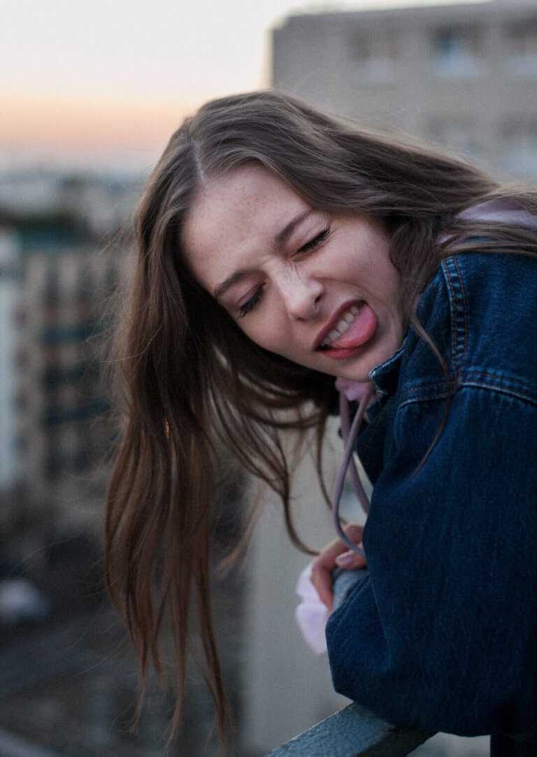 Silly portrait of a young woman in denim jacket sticking her tongue out on a rooftop with a city skyline in the blurred background.