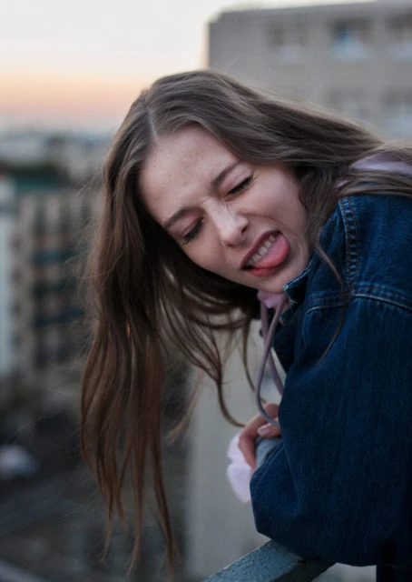 Silly portrait of a young woman in denim jacket sticking her tongue out on a rooftop with a city skyline in the blurred background.