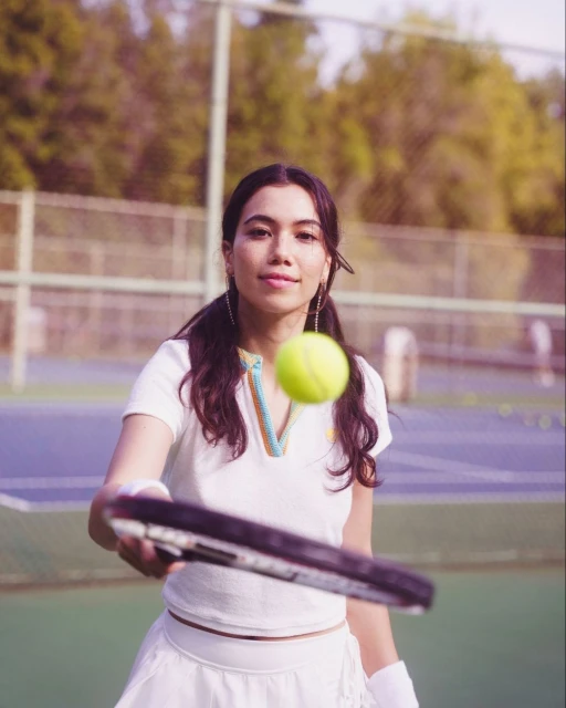 Woman in tennis whites on a blue tennis court holding a racquet with a tennis ball in the air, captured in medium shot in daylight.